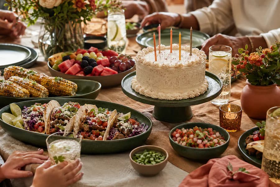 A birthday table with tacos, grilled corn, fruit, drinks, and a candle-topped cake.