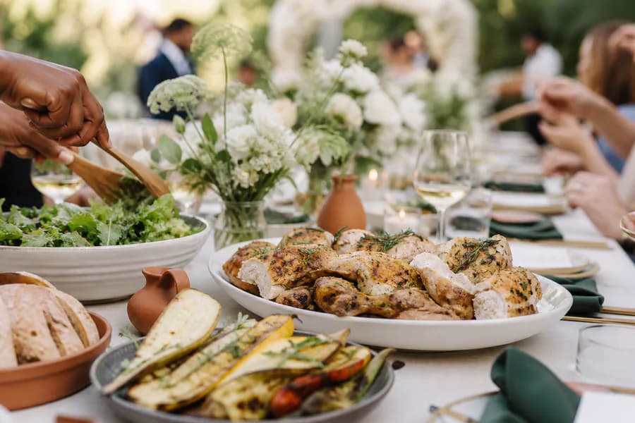 An outdoor wedding reception table with roast chicken, salad, vegetables, and bread.